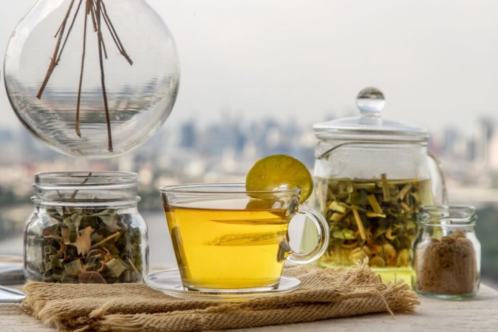 Set-up pe o terasă cu o ceașcă de ceai de lemongrass decorată cu limetă alături de 2 borcane cu lemongrass tocat și uscat