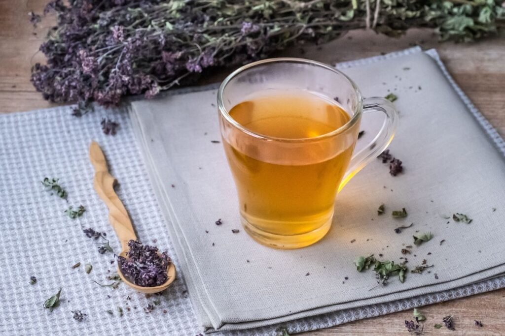 Ceașcă de ceai de oregano pe un caiet, alături de o linguriță cu flori de oregano și un mănunchi de flori.