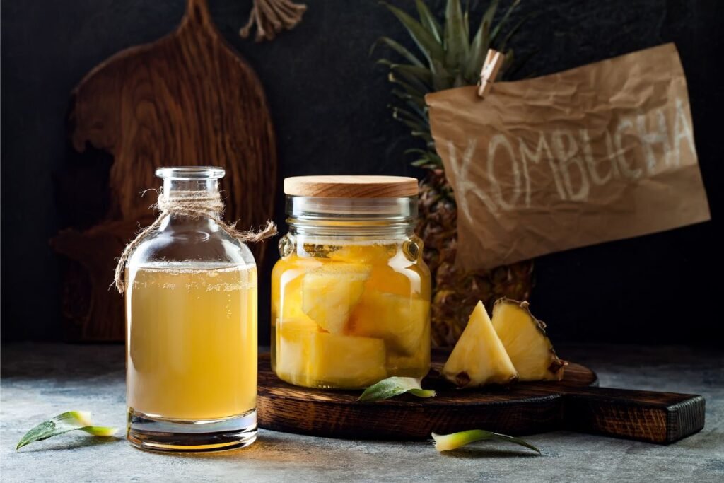 Sticlă artizanala cu ceai de ananas alături de un borcan cu capac de lemn plin cu bucăți de ananas.